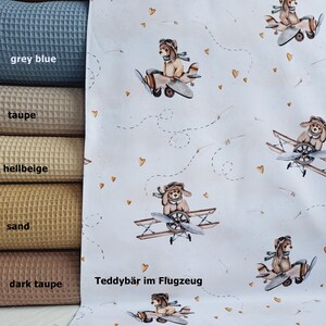 May include: White fabric with a repeating pattern of teddy bears wearing aviator goggles and flying in airplanes. The airplanes have brown propellers and the teddy bears are wearing brown and white striped shirts. The background is white with small gold hearts and dashed lines that look like flight paths. The text "Teddyb&auml;r im Flugzeug" is printed in black at the bottom of the fabric.