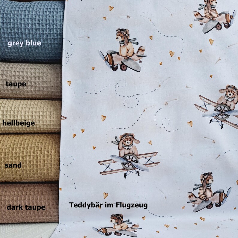 May include: White fabric with a repeating pattern of teddy bears wearing aviator goggles and flying in airplanes. The airplanes have brown propellers and the teddy bears are wearing brown and white striped shirts. The background is white with small gold hearts and dashed lines that look like flight paths. The text "Teddyb&auml;r im Flugzeug" is printed in black at the bottom of the fabric.
