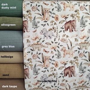 May include: A white fabric with a repeating pattern of prehistoric animals, including a triceratops, a stegosaurus, a saber-toothed cat, and a mammoth. The pattern also includes ferns, flowers, and leaves. The text "Vorgeschichte" is printed in the bottom right corner of the fabric.