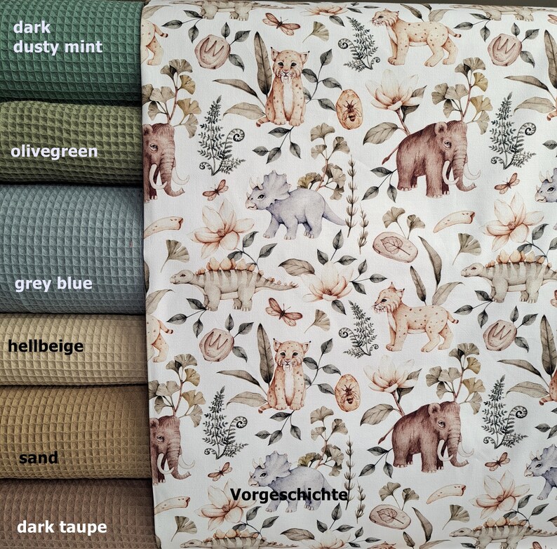 May include: A white fabric with a repeating pattern of prehistoric animals, including a triceratops, a stegosaurus, a saber-toothed cat, and a mammoth. The pattern also includes ferns, flowers, and leaves. The text "Vorgeschichte" is printed in the bottom right corner of the fabric.