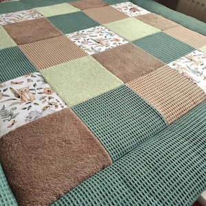 Könnte beinhalten: Eine Patchwork-Decke mit Quadraten aus verschiedenen Texturen und Farben. Die Decke enthält Quadrate aus grünem Waffelgewebe, hellbraunem und gemustertem Stoff mit floralen und tierischen Motiven. Das Gesamtdesign ist eine Mischung aus Texturen und Erdtönen.
