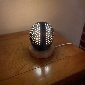 May include: A black, spherical lamp with numerous small, evenly spaced holes, emitting a warm glow. The lamp sits on a round, light-colored wooden base. A white cord extends from the lamp, suggesting it is plugged in. The lamp is on a wooden table.