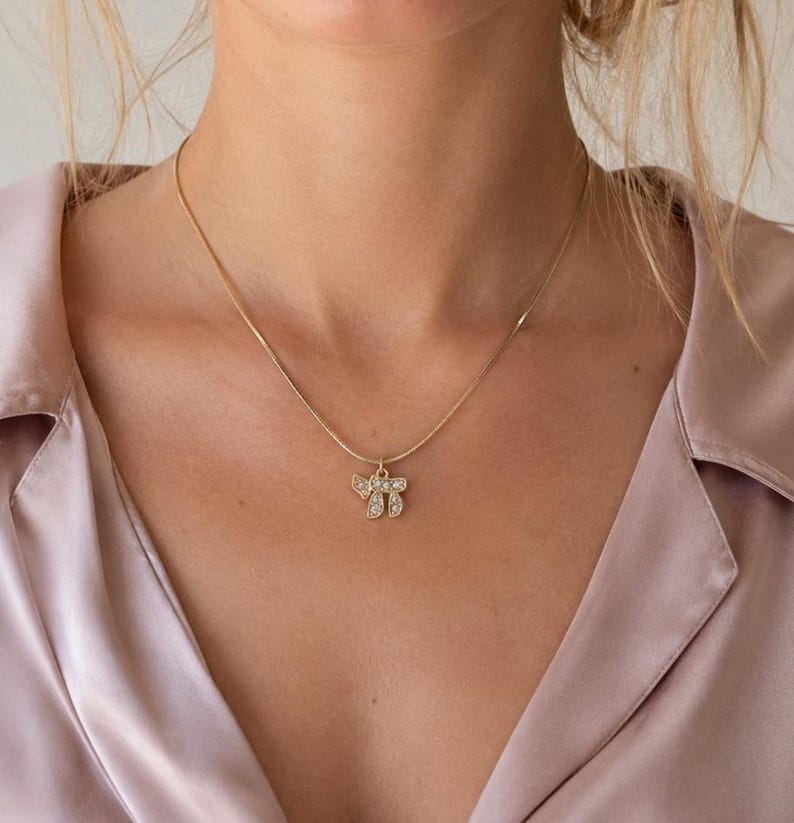 May include: A gold-toned chain necklace featuring a small pendant shaped like the Hebrew letters Chai, which are encrusted with small, clear, sparkling stones. The necklace rests against a light pink satin collared shirt with a visible button placket.