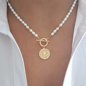 May include: A gold necklace with a pearl chain and a large, round pendant featuring an evil eye design with a white stone in the center. The pendant is surrounded by smaller stones.