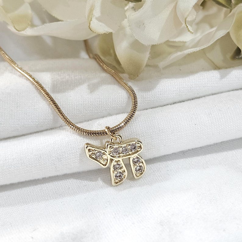 May include: A gold-toned snake chain necklace featuring a pendant shaped like the Hebrew letter Chai. The pendant is embellished with small, clear, sparkling stones set into the metal surface, creating a bright and reflective appearance.