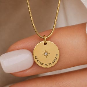 May include: A gold-toned circular pendant necklace featuring a delicate snake chain. The pendant has a scalloped edge, a central starburst motif with a small embedded clear stone, and the engraved coordinates 43.6418 N, 71.4460 W.
