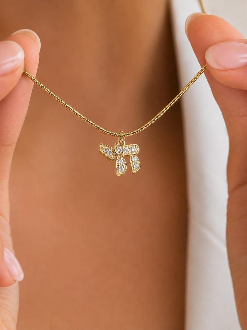 May include: A gold-toned necklace featuring a delicate snake chain and a pendant shaped like the Hebrew letter Chai, encrusted with small, clear, sparkling stones. The pendant hangs from a small circular jump ring attached to the chain.