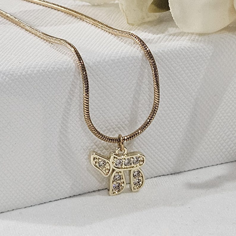 May include: A gold-toned snake chain necklace featuring a pendant shaped like the Hebrew character Chai. The pendant is embellished with small, clear, sparkling stones set into the metal surface, creating a bright and reflective appearance.