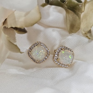 May include: A pair of gold earrings with white opal stones set in a square shape with a halo of smaller clear stones.