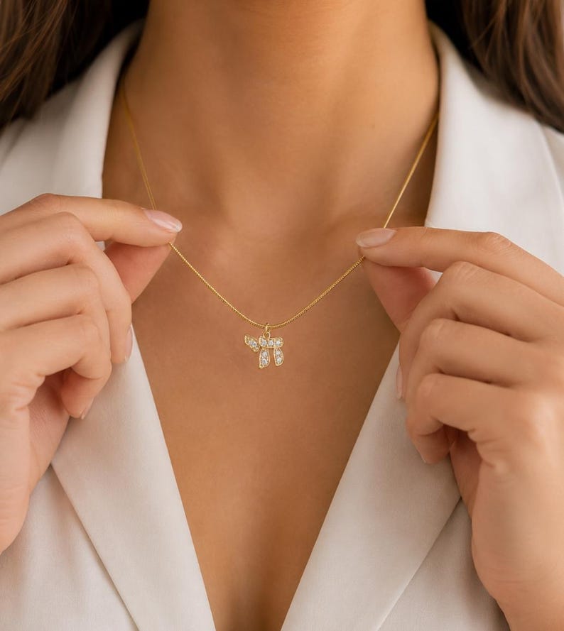 May include: A gold-toned necklace featuring a delicate chain and a small pendant shaped like the Hebrew letter Chai, which is encrusted with small, clear, sparkling stones. The pendant hangs centrally from the thin, polished gold-colored chain.