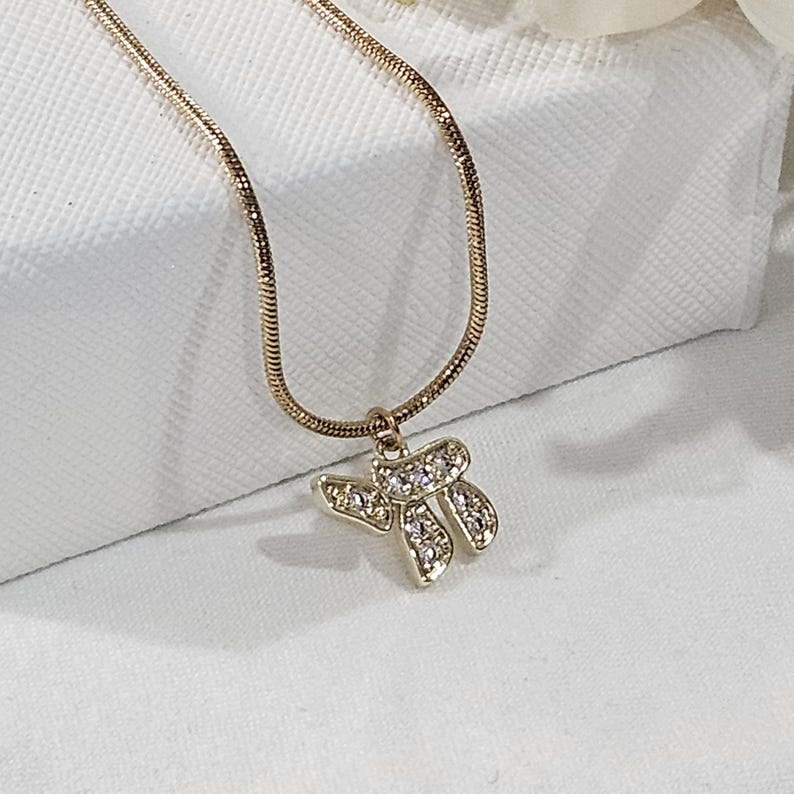 May include: A gold-toned snake chain necklace featuring a pendant shaped like the Hebrew letter Chai. The pendant is embellished with small, clear, sparkling stones set into the metal surface, creating a bright and reflective appearance.