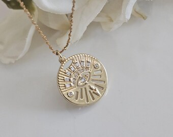 May include: Gold necklace with a round pendant featuring a stylised eye with clear crystal accents. The pendant has a sunburst design around the eye.