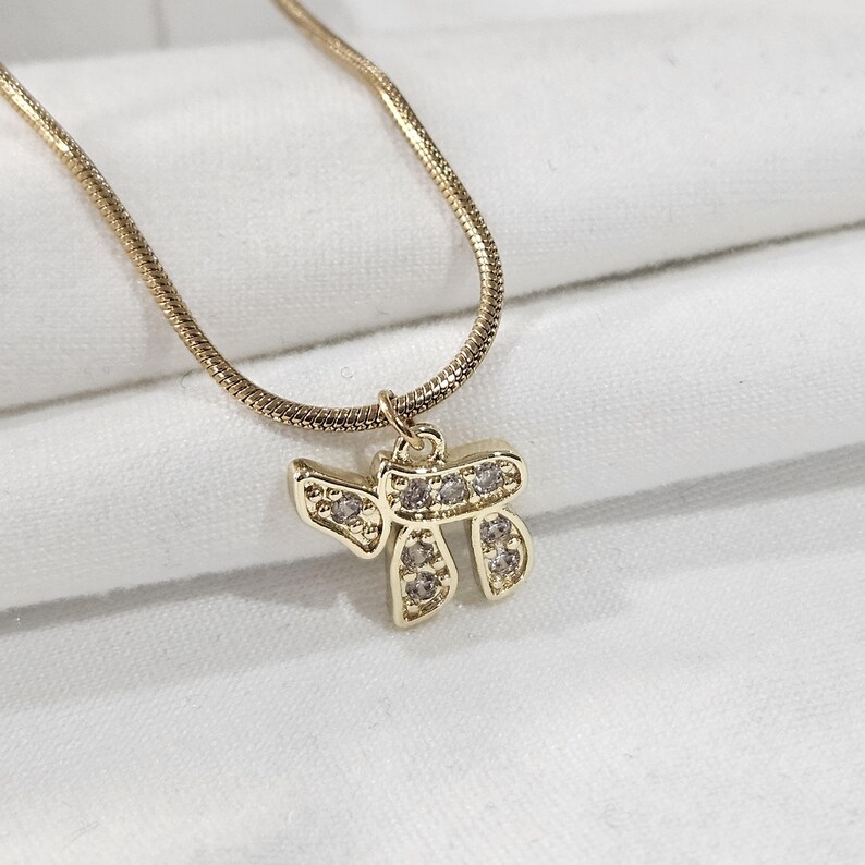 May include: A gold-toned snake chain necklace featuring a pendant shaped like the Hebrew letter Chai. The pendant is embellished with small, clear, sparkling stones set into the metal frame, creating a bright and reflective appearance.