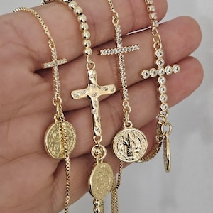 Peut inclure: Trois bracelets en chaîne dorée avec des breloques en forme de croix et des médaillons. Chaque bracelet présente un style de croix différent et un médaillon rond. Les médaillons ont un design en relief.