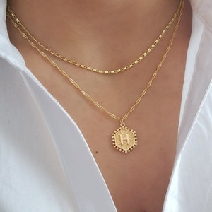 May include: A gold chain necklace with a hexagonal pendant featuring the letter "H" in a gold finish. The pendant is surrounded by small gold beads.