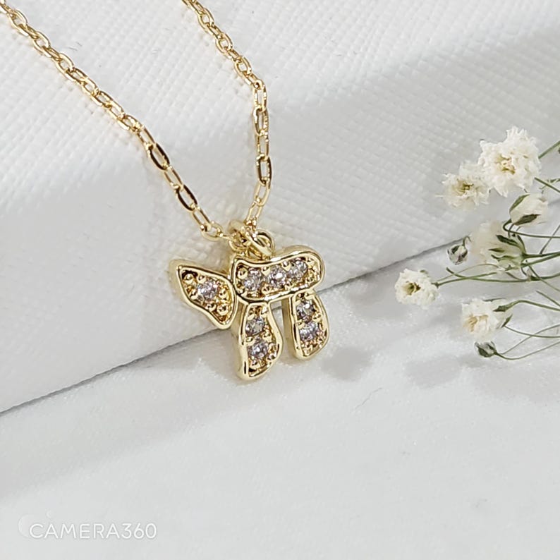 May include: A gold-toned necklace featuring a pendant shaped like the Hebrew letter Chai, encrusted with small clear stones. The pendant hangs from a delicate gold-toned chain, showcasing a polished finish and a reflective, metallic appearance.