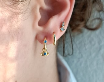May include: Gold hoop earrings with a dangling evil eye charm featuring blue gemstones. The earrings are worn in the earlobe and helix piercing.