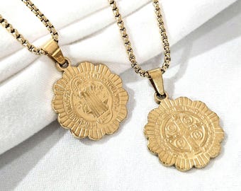 May include: Two gold-colored pendant necklaces. Each necklace features a round pendant with a detailed religious design and a decorative border. The pendants hang from delicate gold chains, set against a white background.