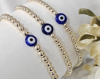 May include: Three beaded bracelets with gold-colored beads and a blue and white evil eye charm. The bracelets are displayed on a white surface, with a soft focus background.