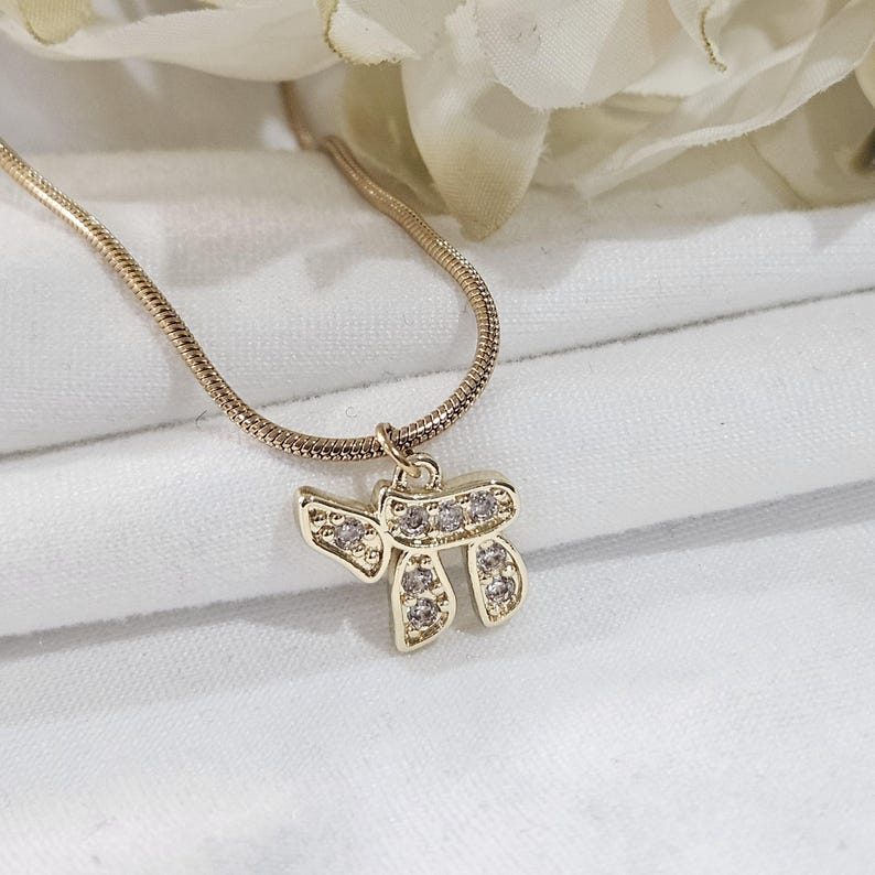May include: A gold-toned necklace featuring a delicate snake chain and a pendant shaped like the Hebrew letter Chai. The pendant is embellished with small, clear, sparkling stones set into the metal, creating a bright and reflective appearance.
