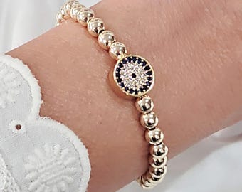 May include: Gold beaded bracelet featuring a central charm with a blue and white design. The bracelet is made of small, round, gold-colored beads. The charm has a circular shape with a dark blue border and a white center.