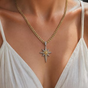 May include: A gold chain necklace featuring a starburst pendant. The pendant has a central gold star element surrounded by silver-toned rays encrusted with small clear stones. The necklace rests against a white garment, showcasing the metal contrast.