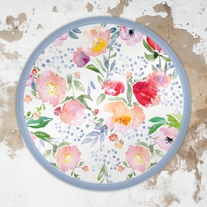 May include: A round, blue-framed wall decor with a watercolour floral pattern. The design features pink, red, and yellow flowers with green leaves on a white background.