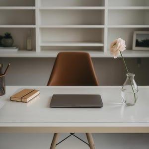 May include: A modern workspace featuring a white desk with a closed laptop, a tan notebook, and a clear glass vase holding a single light pink flower. A brown chair sits in the background, with white shelves above.