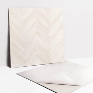May include: A square adhesive tile featuring a light beige and off-white herringbone wood grain pattern. The tile has a textured surface and a peel-off backing layer partially detached at the bottom corner, revealing a smooth white adhesive side.