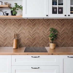 May include: A kitchen backsplash featuring a brown wood-look herringbone pattern tile design. The backsplash is positioned between white upper cabinets and a wooden countertop, with a black stovetop, a wooden knife block, and a small potted plant.