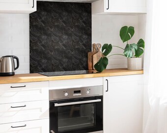 May include: A kitchen splashback featuring a dark black marble pattern with subtle white veining, installed behind a stainless steel electric hob and oven, set against white cabinetry and a light wood worktop with a green potted plant.