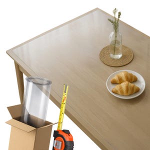 May include: A light wood dining table with a clear plastic table protector. A measuring tape is next to a cardboard box containing a roll of plastic. A small vase with greenery and a plate with two croissants are on the table.