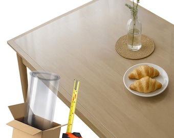 May include: A light wood dining table with a clear plastic table protector. A measuring tape is next to a cardboard box containing a roll of plastic. A small vase with greenery and a plate with two croissants are on the table.