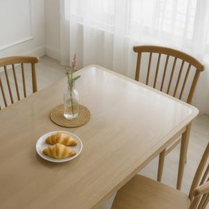 Könnte beinhalten: Ein Esstisch aus hellem Holz mit zwei Croissants auf einem weißen Teller. Eine klare Glasvase mit einem Blütenzweig steht auf einem gewebten Untersetzer. Drei Holzstühle umgeben den Tisch und schaffen eine gemütliche Essumgebung.