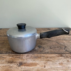 May include: A silver-coloured aluminium saucepan with a matching lid. The lid features a small, round black knob. The saucepan has a long, textured black handle with a metal loop at the end for hanging. The exterior surface shows light wear.