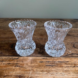 May include: Two clear, cut-glass bud vases with a detailed, textured pattern. The vases have a flared top and a rounded base, sitting on a wooden surface. The glass reflects light, creating a sparkling effect. These decorative vases are ideal for small floral arrangements.