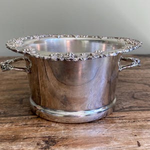 May include: A polished silver-toned metal wine cooler or ice bucket with a wide, flared rim featuring a detailed embossed grape and vine leaf pattern. The vessel has two curved side handles and a smooth, cylindrical body with a rounded base.