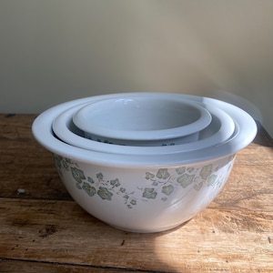 May include: A set of three white ceramic mixing bowls. The bowls nest inside each other. The largest bowl has a floral pattern in shades of green and grey. The bowls are on a wooden surface.