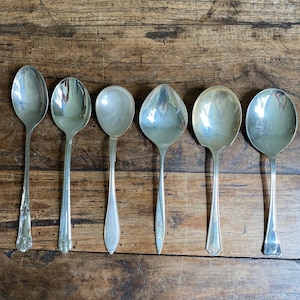 Peut inclure: Un ensemble de six cuillères en métal argenté disposées en rangée. Chaque cuillère présente un motif de manche distinct, allant de motifs floraux ornés à des formes lisses et effilées, avec des bols de tailles et de formes variées.