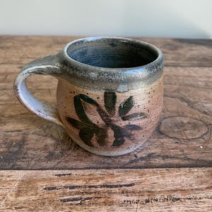 Peut inclure: Une tasse en céramique ronde au corps beige et brun moucheté, ornée d'un motif floral peint en vert foncé sur le devant. La tasse possède une anse épaisse et texturée ainsi qu'un bord métallique lisse qui se fond dans un intérieur bleu profond.