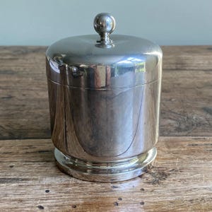 May include: A silver-toned, cylindrical container with a lid and a small, spherical knob. The container has a wide base and a smooth, reflective surface. It is likely a decorative storage jar or a small canister, suitable for various uses.