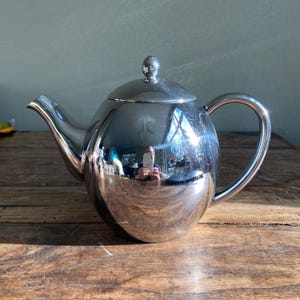 May include: A polished silver teapot with a spherical body, a curved spout, and a loop handle. The lid is topped with a small, round knob. The teapot reflects the surrounding environment, including the wooden surface it sits on.