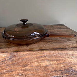 May include: A vintage brown glass skillet with a matching lid. The skillet has a long handle and a rounded shape. The lid has a small, round knob. The glass has a slightly transparent quality. The skillet is resting on a wooden surface. The dimensions are approximately 25 cm in diameter.