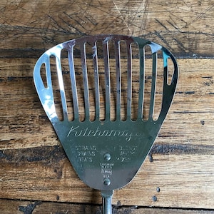 May include: Vintage chrome-plated kitchen utensil with a slotted design. The utensil is marked "Kitchamajig" and features text for straining, draining, and mixing. Made in the USA.