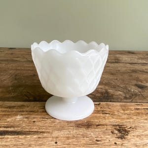 Peut inclure: Un bol à pied blanc de style vintage avec un bord festonné et un motif en losange. Le bol est en verre opaque et repose sur une base ronde. Le bol est probablement utilisé pour servir des desserts ou exposer de petits objets.