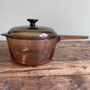 May include: A brown glass saucepan with a lid and a handle. The saucepan is sitting on a wooden surface.
