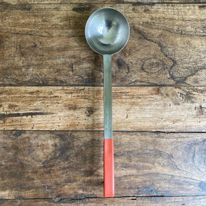 May include: A stainless steel ladle with a round bowl and a long, flat handle. The handle has a red grip at the end. The ladle is designed for serving soups, sauces, or other liquids. The kitchen utensil is shown against a wooden background.