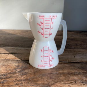 May include: White plastic measuring jug with red markings. The jug has a handle and measures up to 8 ounces or 1 cup.