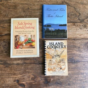 May include: Three vintage cookbooks on a wooden surface. Titles include "Salt Spring Island Cooking", "Tastes and Tales of Thetis Island", and "Island Cookery". The cookbooks feature images of food and landscapes.