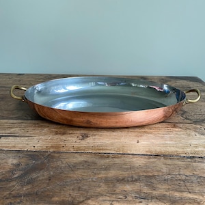 May include: An oval copper pan with brass handles. The interior is silver-coloured, and the exterior is a warm copper tone. The pan is on a wooden surface. This kitchenware item is designed for cooking and serving.